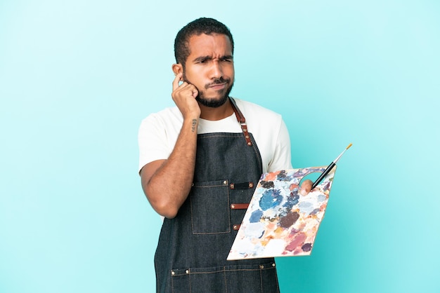 Homem latino jovem artista segurando uma paleta isolada em fundo azul frustrado e cobrindo os ouvidos