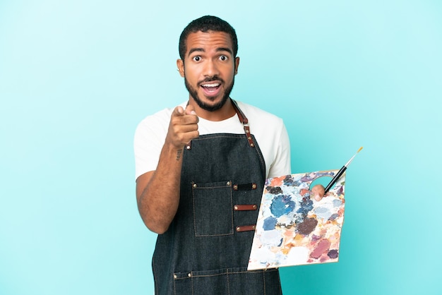 Homem latino jovem artista segurando uma paleta isolada em fundo azul surpreso e apontando para a frente