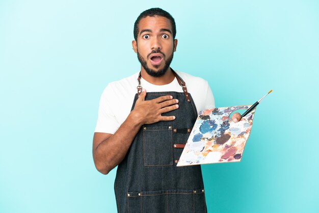 Homem latino jovem artista segurando uma paleta isolada em fundo azul surpreso e chocado ao olhar para a direita