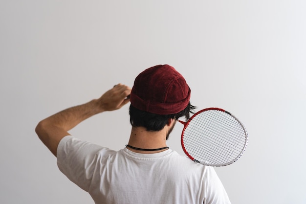 Homem latino jovem hippie com barba virando as costas com um gorro segurando uma raquete de badminton com fundo branco Foto artística