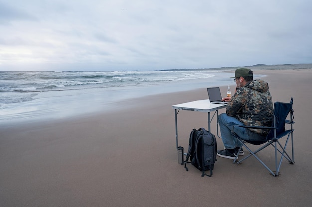 Homem latino maduro sentado na praia trabalhando com seu notebook em uma mesa portátil