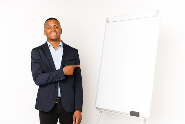 Homem latino novo com um quadro branco em uma parede branca que sorri e que aponta de lado, mostrando algo no espaço em branco.