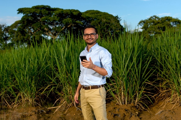 Homem latino olhando diretamente para a câmera segurando seu smartphone e posando em frente a uma plantação de cana-de-açúcar