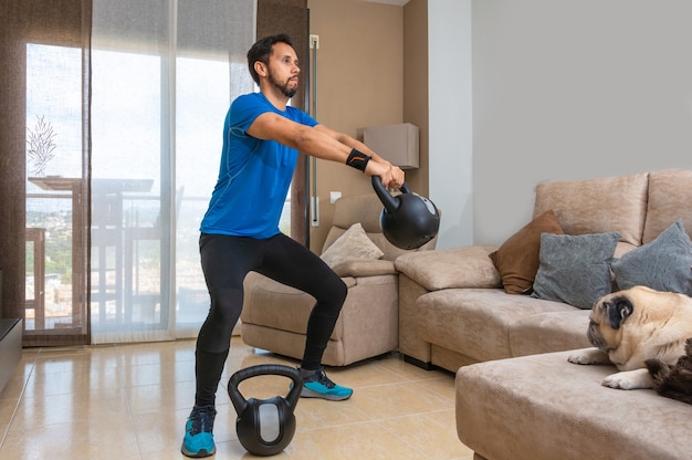 Homem latino realizando um treino de cross fit em casa com um par de kettlebells.