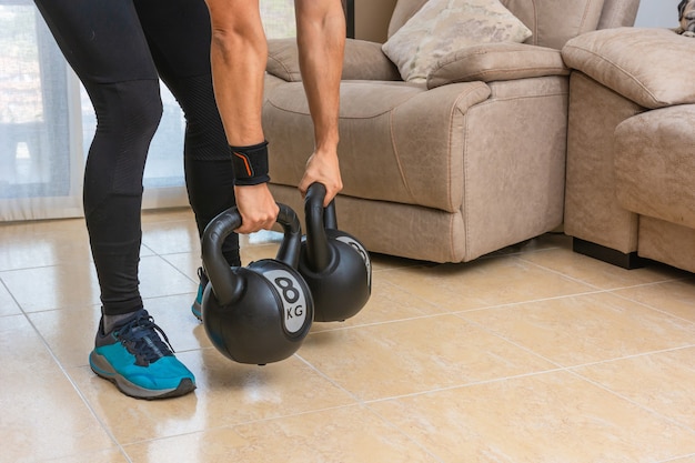 Homem latino realizando um treino de cross fit em casa com um par de kettlebells.