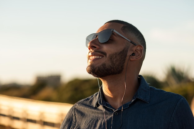 Homem latino usando fones de ouvido para ouvir música ou podcasts motivacionais e apreciando o pôr do sol. Luz quente natural.
