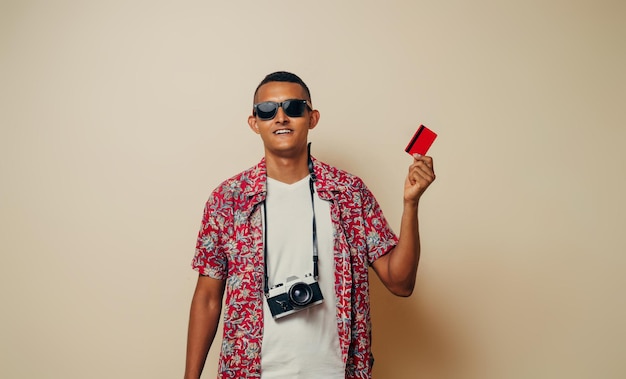 Homem latinx jovem turista com óculos de sol segurando um cartão de crédito vermelho olhando para o conceito de verão da câmera