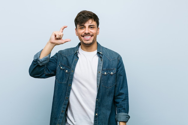 Homem legal hispânico jovem segurando algo pouco com o dedo indicador, sorrindo e confiante.