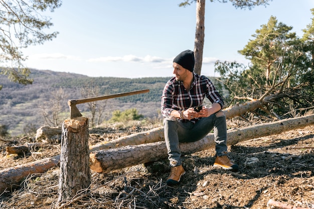 Homem lenhador sentado em um tronco de madeira ao lado de seu machado olhando para o celular