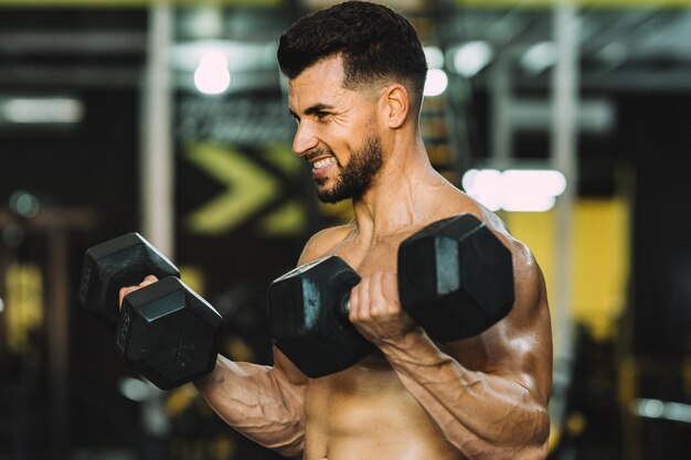 Foto homem levantando halteres metálicos em uma academia com expressão de esforço