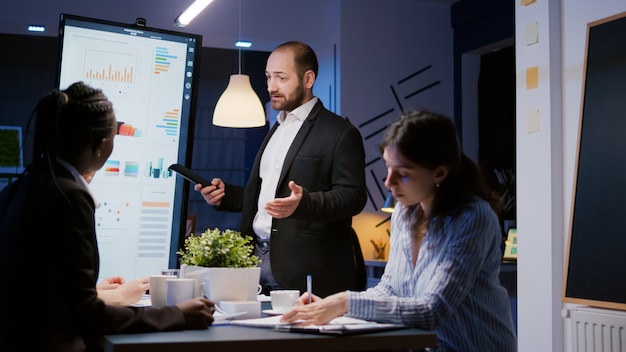 Homem líder focado explicando o projeto de gerenciamento usando monitor trabalhando na sala de reuniões da empresa tarde da noite