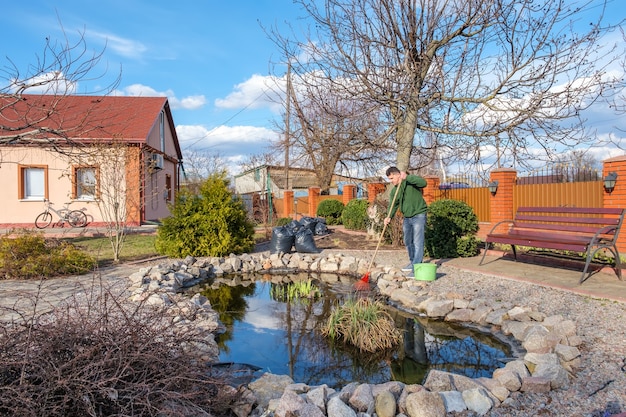 Homem limpa um lago de jardim com um ancinho de plantas aquáticas e folhas caindo