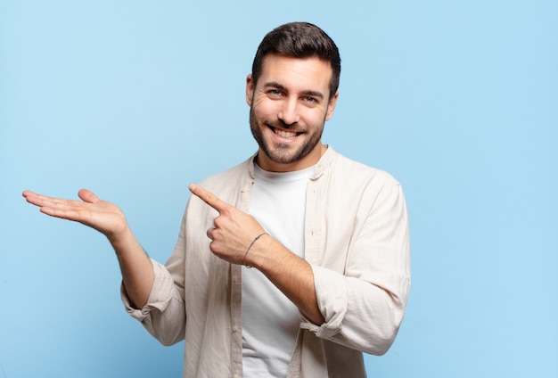 Homem loiro adulto bonito sorrindo alegremente e apontando para copiar o espaço na palma da mão ao lado, mostrando ou anunciando um objeto