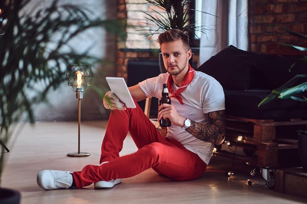 Homem loiro atraente com tatuagens nos braços, vestido com uma calça vermelha de Natal usando tablet PC em uma sala com interior loft.