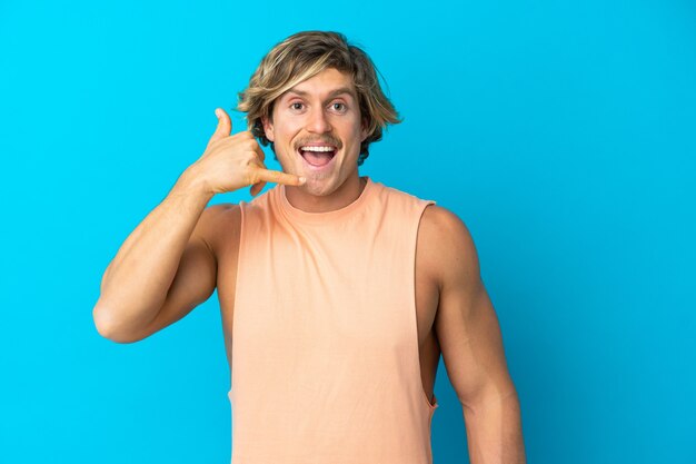 Homem loiro bonito isolado em azul fazendo gesto de telefone