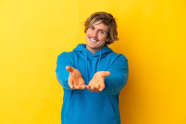 Homem loiro bonito isolado na parede amarela segurando copyspace imaginário na palma da mão para inserir um anúncio