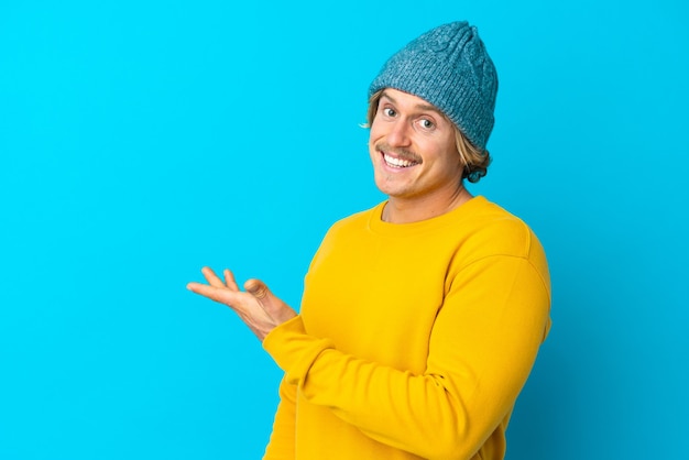 Homem loiro bonito isolado na parede azul apresentando uma ideia enquanto olha sorrindo para