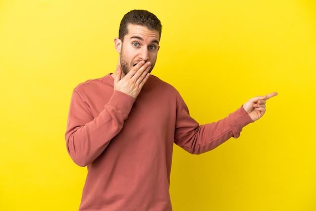 Homem loiro bonito sobre fundo amarelo isolado com expressão de surpresa apontando para o lado