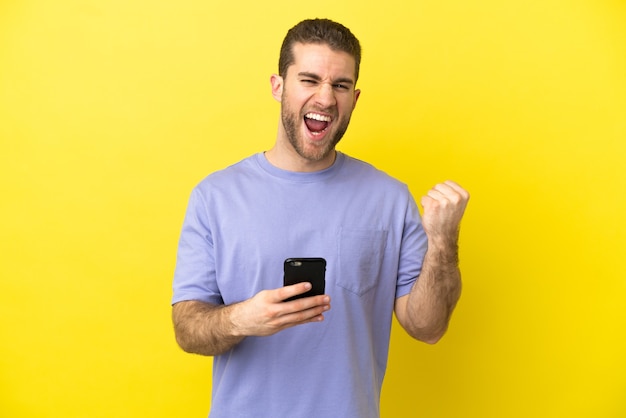 Homem loiro bonito sobre fundo amarelo isolado com o telefone em posição de vitória