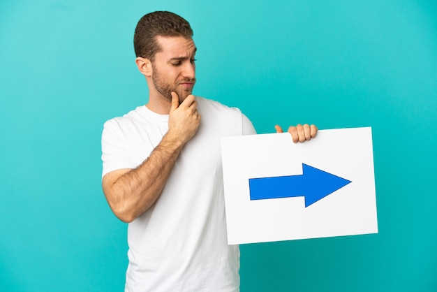 Homem loiro bonito sobre um fundo azul isolado, segurando um cartaz com o símbolo de seta e pensando