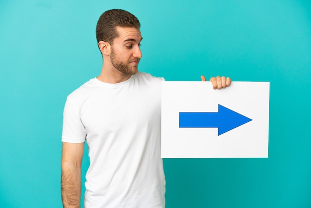 Homem loiro bonito sobre um fundo azul isolado segurando um cartaz com o símbolo de seta