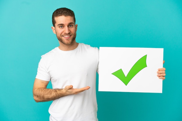 Foto homem loiro bonito sobre um fundo azul isolado segurando um cartaz com o texto ícone de marca de seleção verde e apontando-o