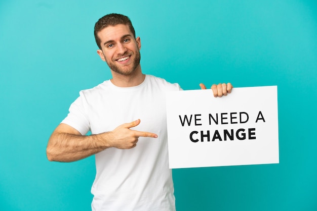 Homem loiro bonito sobre um fundo azul isolado segurando um cartaz com o texto Precisamos de uma mudança e apontando-o