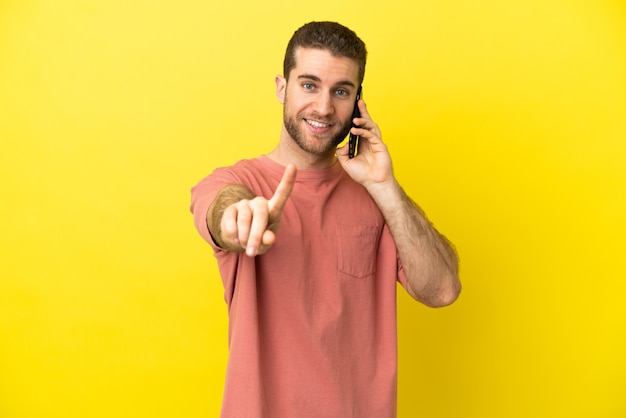 Homem loiro bonito usando telefone celular sobre fundo isolado, mostrando e levantando um dedo