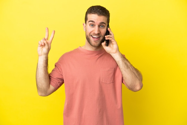 Homem loiro bonito usando telefone celular sobre fundo isolado, sorrindo e mostrando sinal de vitória