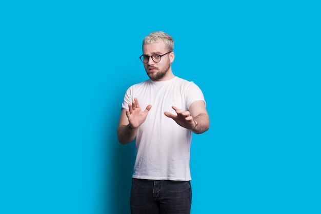 Homem loiro com barba gesticulando com medo enquanto olha pelos óculos e posa em um fundo azul