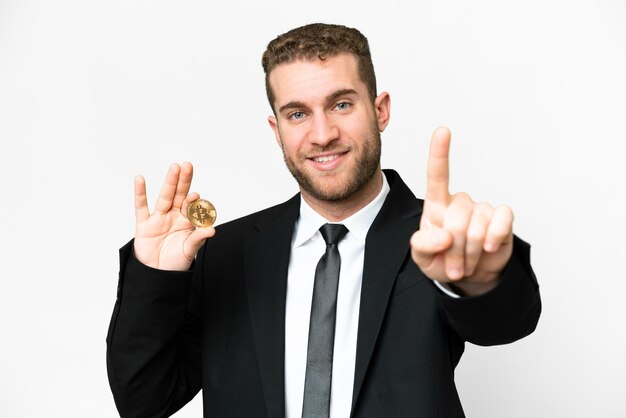 Homem loiro de negócios jovem segurando um Bitcoin sobre fundo branco isolado mostrando e levantando um dedo