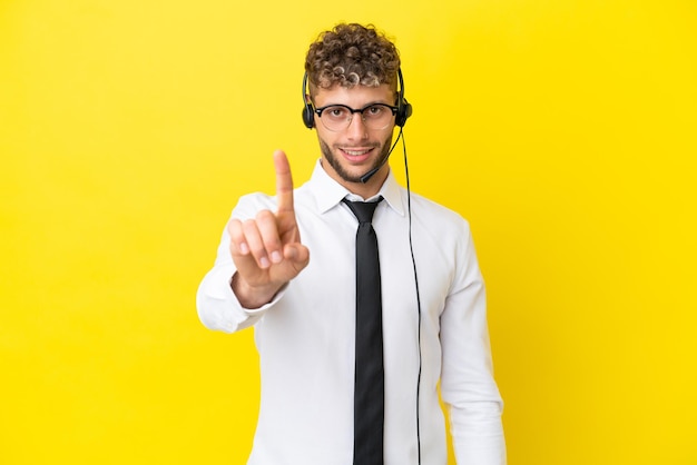 Homem loiro de telemarketing trabalhando com um fone de ouvido isolado em fundo amarelo mostrando e levantando um dedo