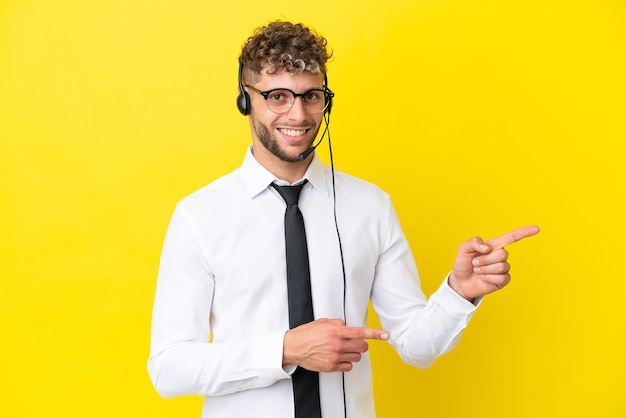 Homem loiro de telemarketing trabalhando com um fone de ouvido isolado em um fundo amarelo, apontando o dedo para o lado e apresentando um produto