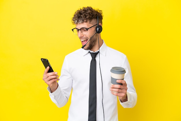 Homem loiro de telemarketing trabalhando com um fone de ouvido isolado em um fundo amarelo, segurando um café para levar e um celular