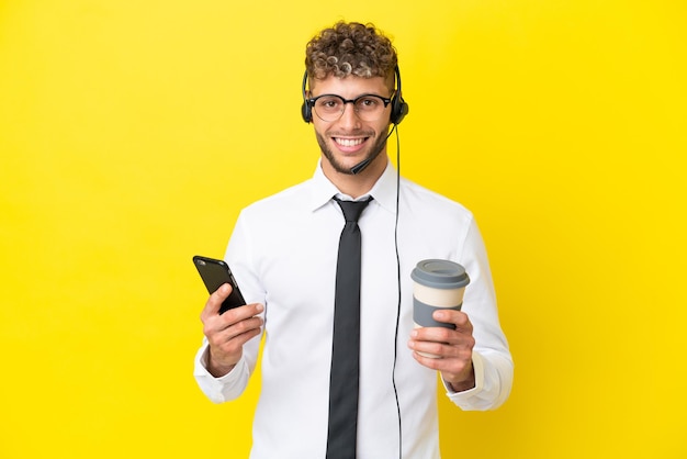 Homem loiro de telemarketing trabalhando com um fone de ouvido isolado em um fundo amarelo, segurando um café para levar e um celular