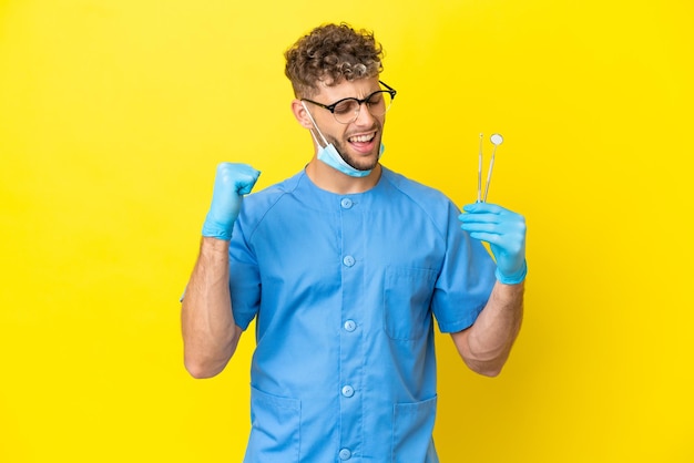 Homem loiro dentista segurando ferramentas isoladas no fundo comemorando uma vitória