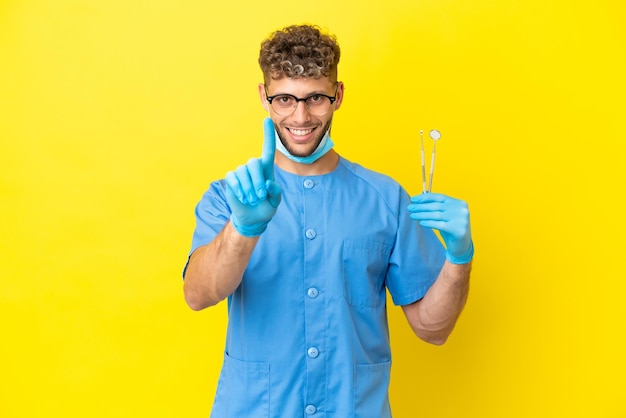 Homem loiro dentista segurando ferramentas isoladas no fundo mostrando e levantando um dedo