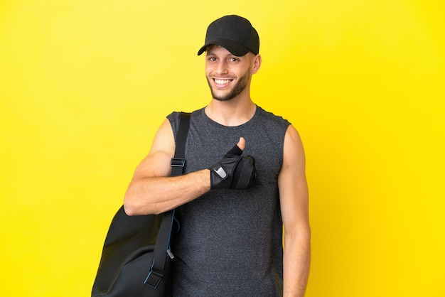 Homem loiro jovem esportivo com bolsa esportiva isolada em fundo amarelo, dando um polegar para cima gesto