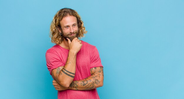 Homem loiro sorrindo com uma expressão feliz e confiante com a mão no queixo, pensando e olhando para o lado