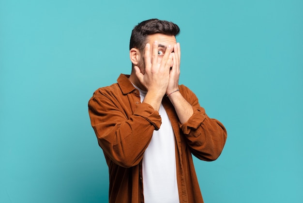 Foto homem louro adulto bonito cobrindo o rosto com as mãos, espiando por entre os dedos com expressão de surpresa e olhando para o lado