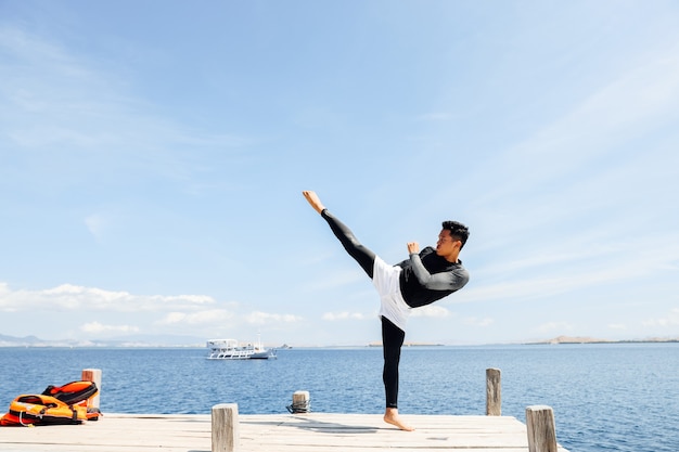 Homem lutador asiático com poses de chute de taekwondo com o mar ao fundo