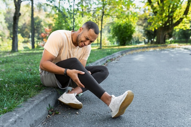 Homem machucou a perna enquanto fazia jogging afro-americano sentado no chão massageando músculos doloridos com