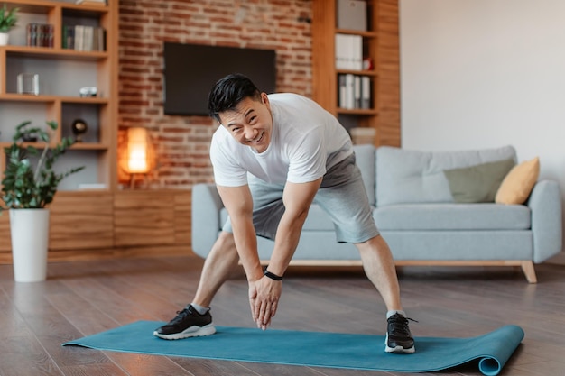 Homem maduro asiático ativo fazendo exercícios esticando os músculos das pernas em casa no tapete de fitness espaço de cópia de comprimento total