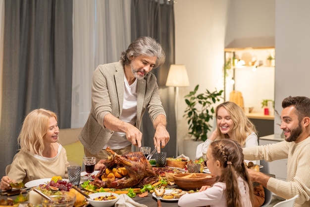 Homem maduro barbudo cortando peru assado caseiro no centro da mesa festiva, enquanto sua família olha para ele no jantar de Ação de Graças