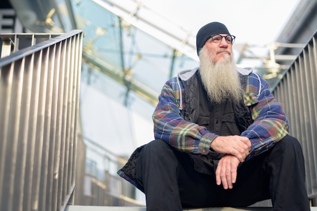 Homem maduro barbudo hippie pensando e sentado na passarela ao ar livre