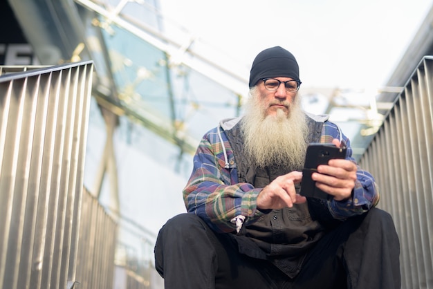 Homem maduro barbudo hippie usando o telefone enquanto está sentado em uma passarela na cidade