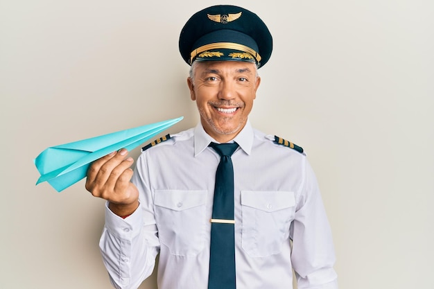 Homem maduro bonito de meia-idade vestindo uniforme de piloto de avião segurando avião de papel olhando positivo e feliz em pé e sorrindo com um sorriso confiante mostrando os dentes