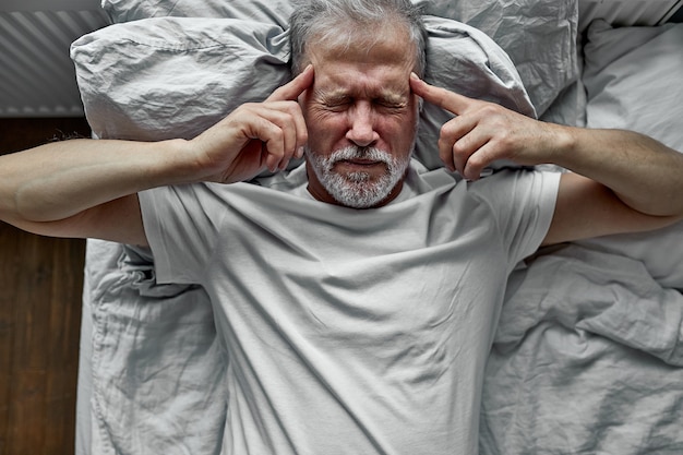 Homem maduro com os olhos fechados, deitado na cama, tocando as têmporas de perto, homem idoso cansado com dor de cabeça ou enxaqueca, mal-estar, insônia e falta de sono