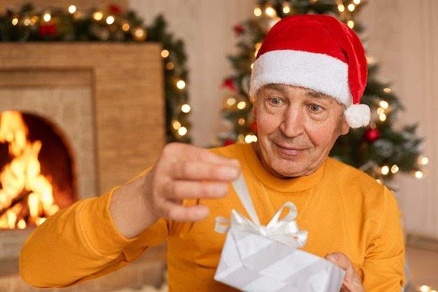 Homem maduro comemorando o natal em casa, abrindo a caixa de presente com fita, sendo surpreendido, sendo fotografado na sala de estar com lareira.
