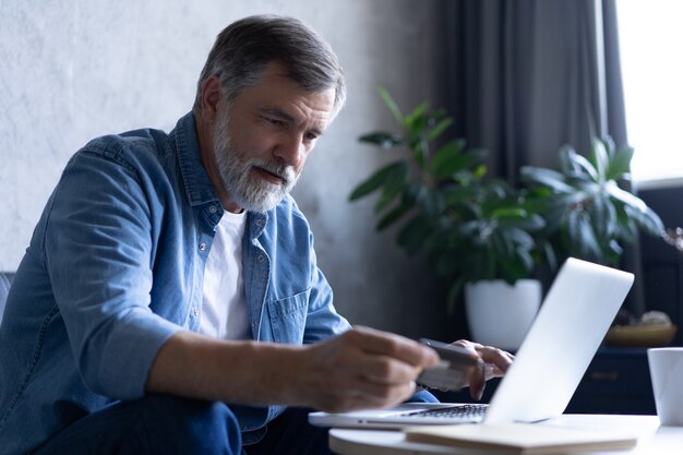 Homem maduro, compras online no laptop, usando o cartão de crédito em casa.